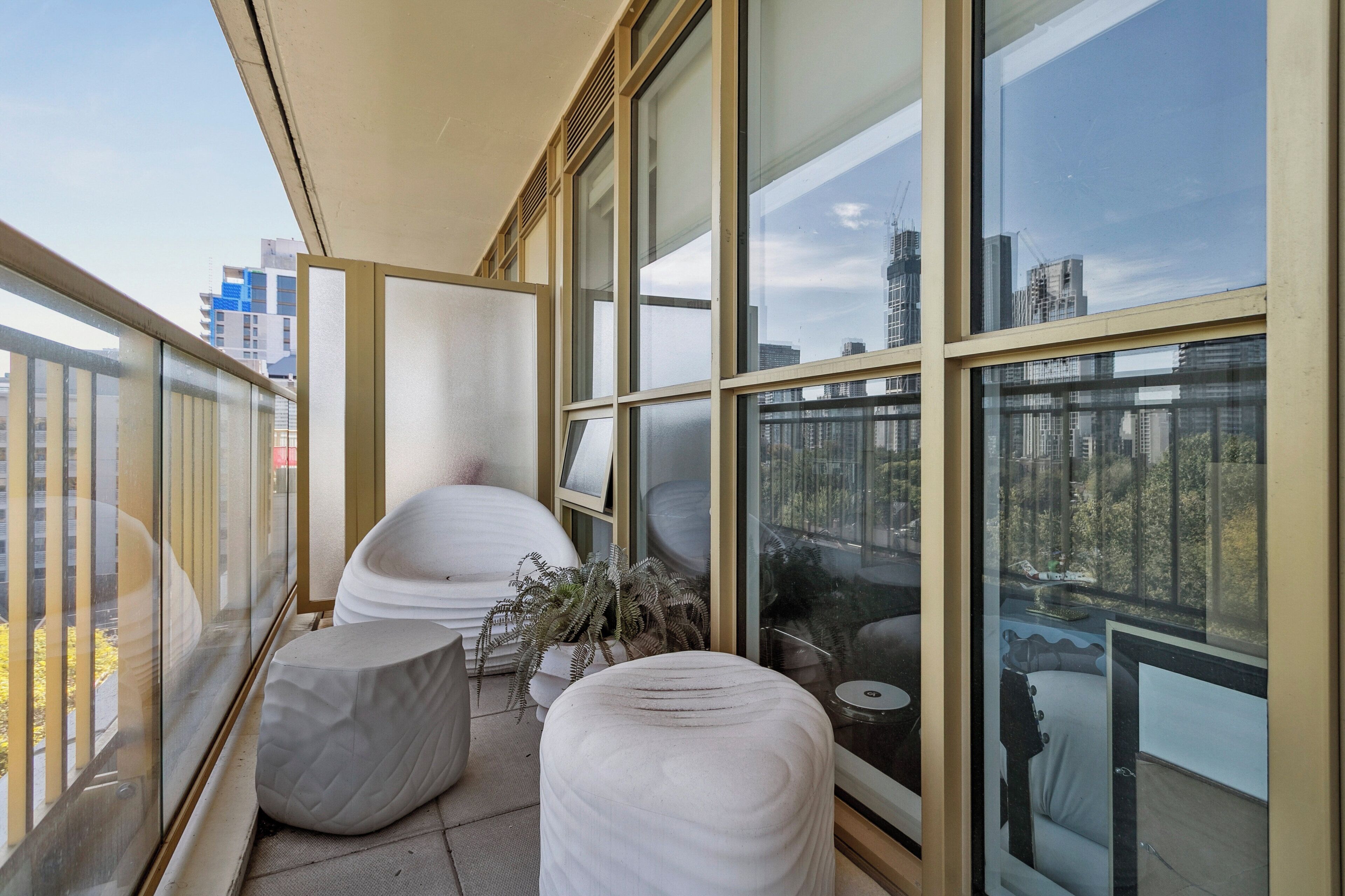 A modern balcony with large windows, white cushioned seating, and a potted plant.