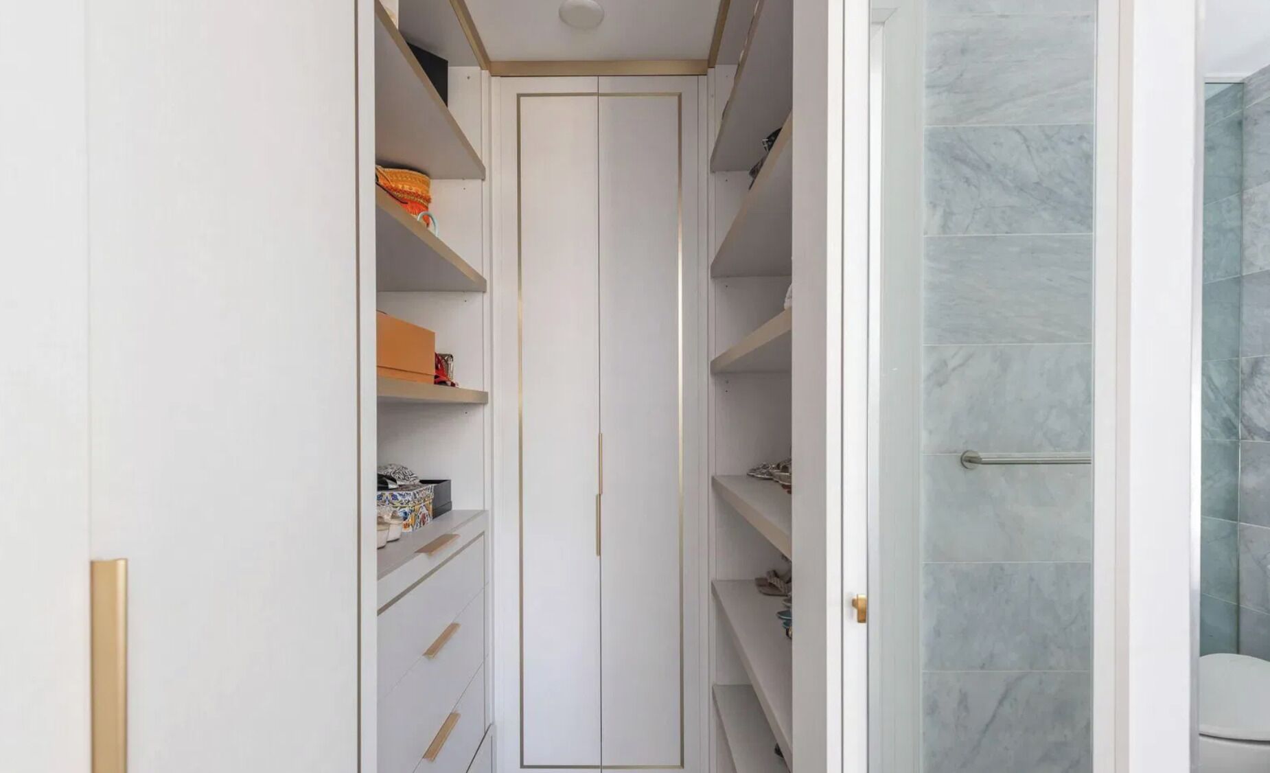 A narrow walk-in closet with shelves and drawers, leading to a bathroom with a glass shower enclosure.