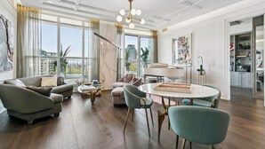 Een ruime woonkamer met een moderne kroonluchter, een eethoek met een ronde tafel en uitzicht op de stad door grote ramen.