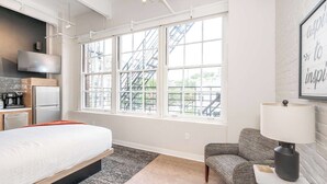 A modern hotel room with a bed, a desk, a chair, a lamp, and a large window with a view of a building. - The 333 Suites 204 Stay for 2 (Cincinnati)