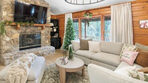 A cozy living room with a stone fireplace, a flat-screen TV, a Christmas tree, and a comfortable sofa. - Stay at Wild & Blue, Log Home Retreat, Game Room w/ Arcade (White Bluff)