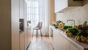 Una cucina piccola con una sola sedia, una finestra con tende leggere e un piano di lavoro con prodotti freschi e bicchieri.