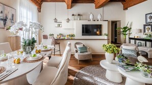 Un ampio soggiorno con soffitto in legno, una zona pranzo con tavolo apparecchiato per quattro persone e un angolo cottura con televisore e oggetti decorativi.