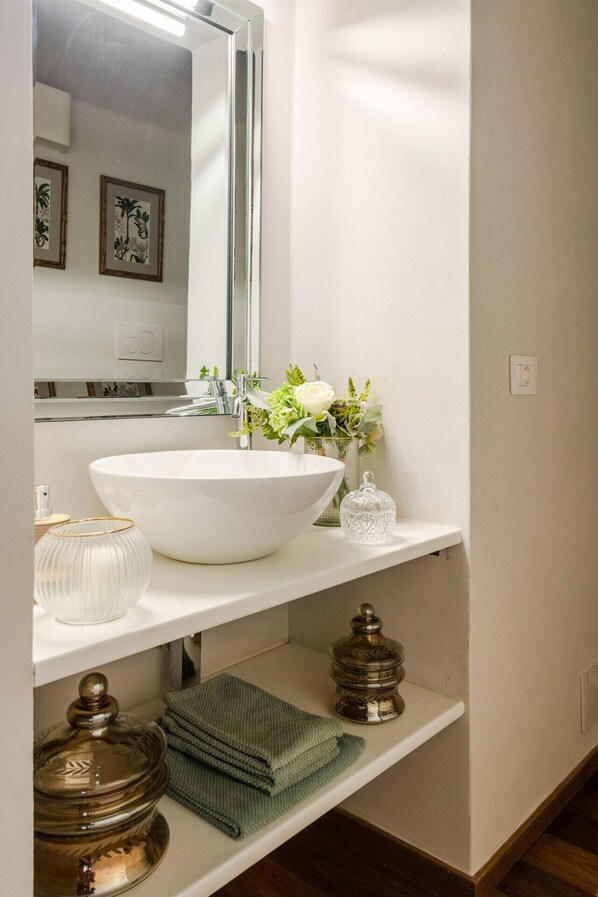Ein Badezimmer mit einem weißen Waschbecken, einem Spiegel, einer Vase mit Blumen und einem Regal mit Handtüchern und einem Glas.