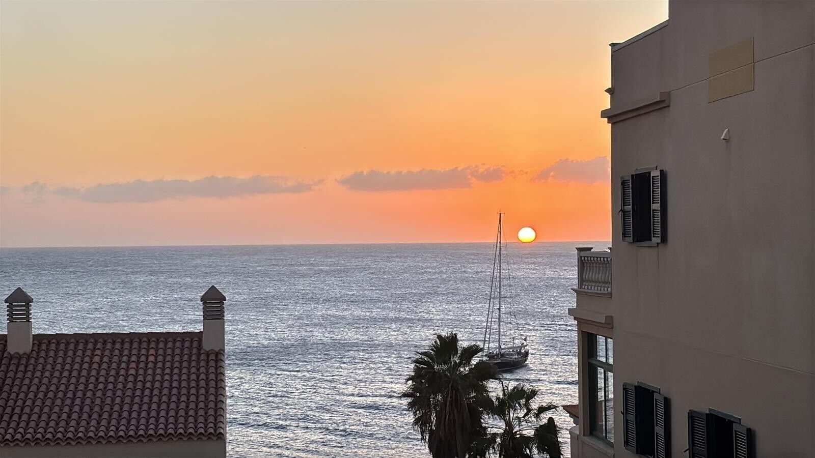 A sunset view from a balcony overlooking the sea, with a sailboat visible in the distance.