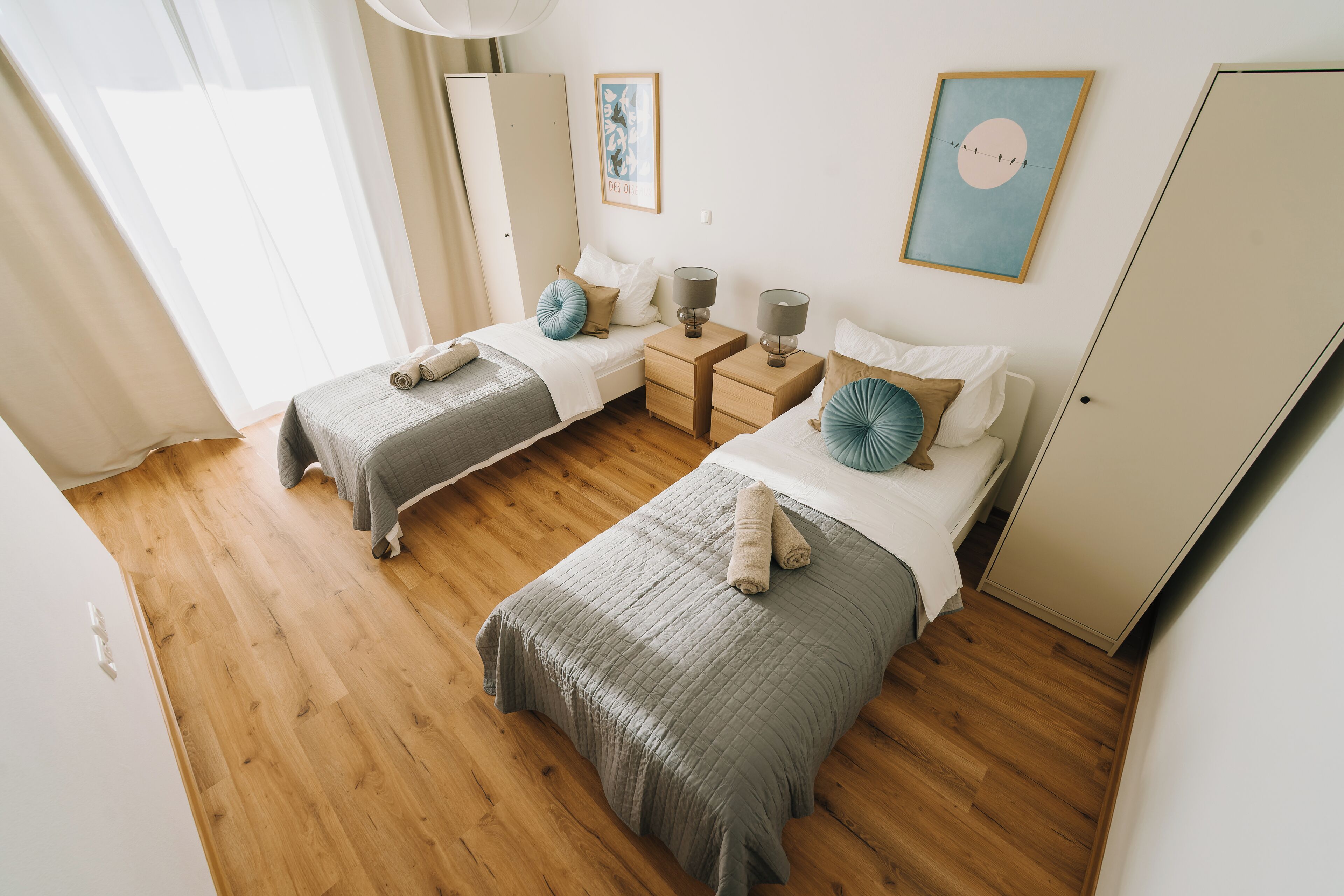 Un dormitorio con dos camas, suelo de madera, mesitas de noche con lámparas y un armario.