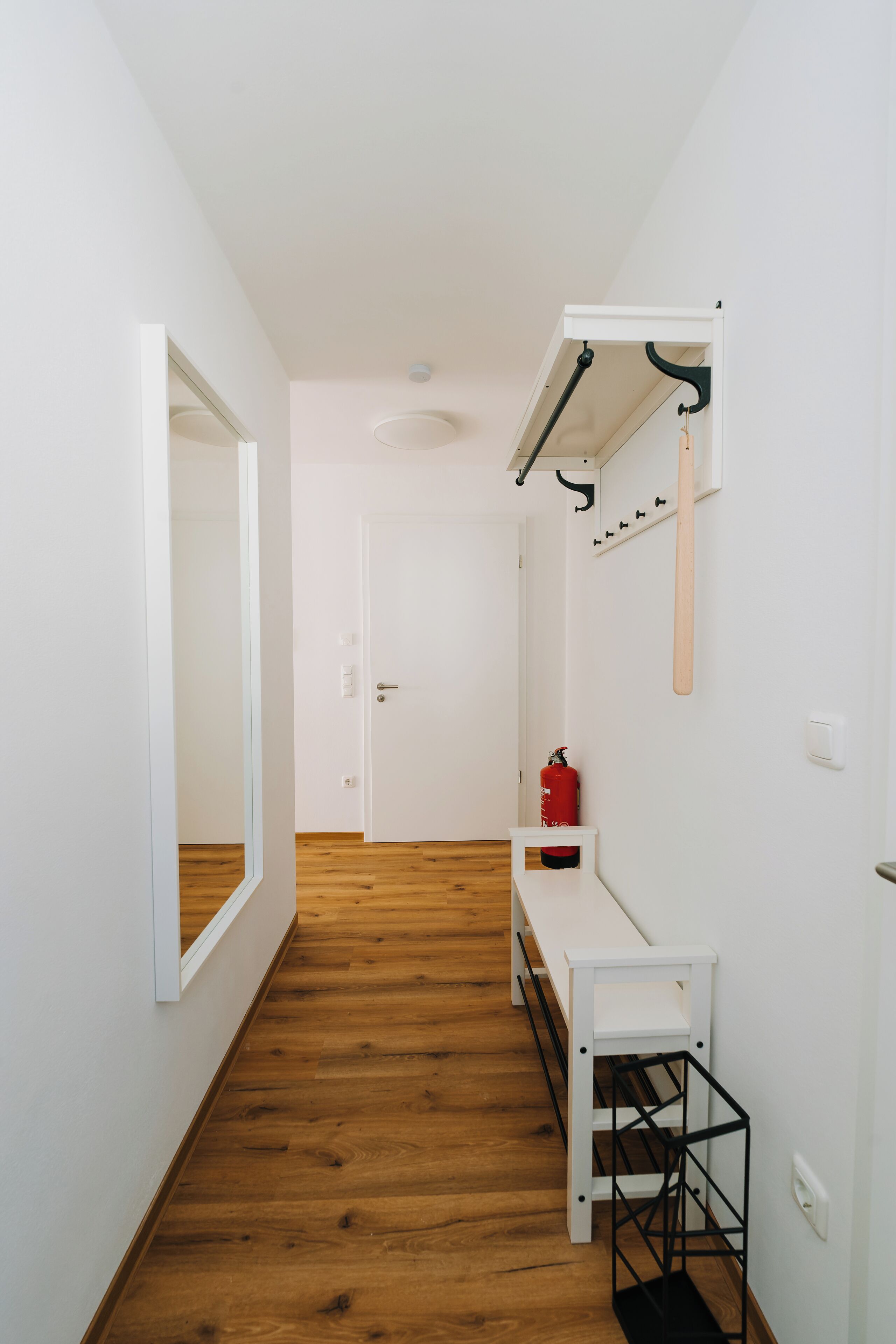 A hallway with wooden flooring, a white bench, a fire extinguisher, and a mirror on the wall.