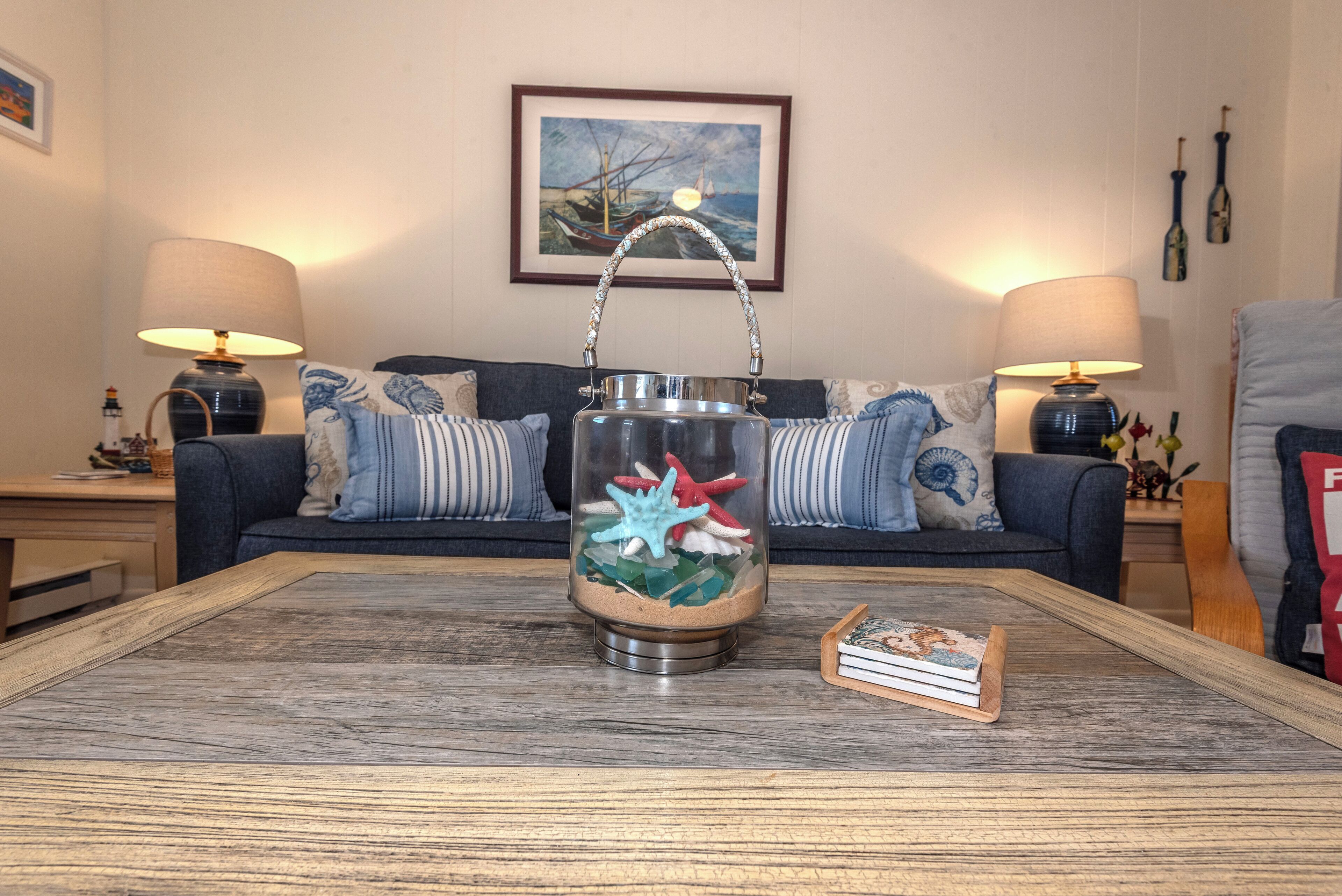 A living room with a coffee table, a glass-top table lamp, a framed painting, and a sofa with striped cushions.