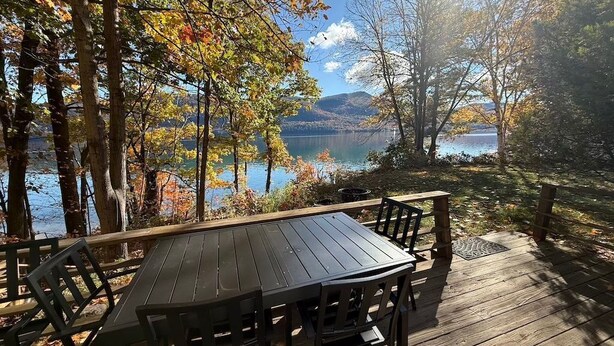 A wooden deck with a table and chairs overlooking a lake with autumn foliage. - Silver Bay Cabin-last minute availability 6/18 (Hague)