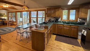 A spacious kitchen with wooden cabinets, a dining area with a table and chairs, and large windows offering a view of the outdoors. - Silver Bay Cabin-last minute availability 6/18 (Hague)