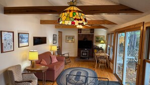 A cozy living room with a fireplace, wooden beams, a dining area, and large windows offering a view of the outdoors. - Silver Bay Cabin-last minute availability 6/18 (Hague)