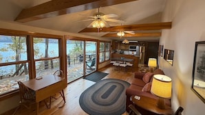 A spacious living room with a large wooden dining table, a sofa, and a fireplace. There are large windows offering a view of a lake. - Silver Bay Cabin-last minute availability 6/18 (Hague)