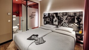 A modern hotel room with a large bed, a headboard with artwork, a red accent wall, and a view of the city through the window. - LÉGÈRE EXPRESS Hotel Leipzig - Apartment with kitchenette & breakfast optional (Leipzig)