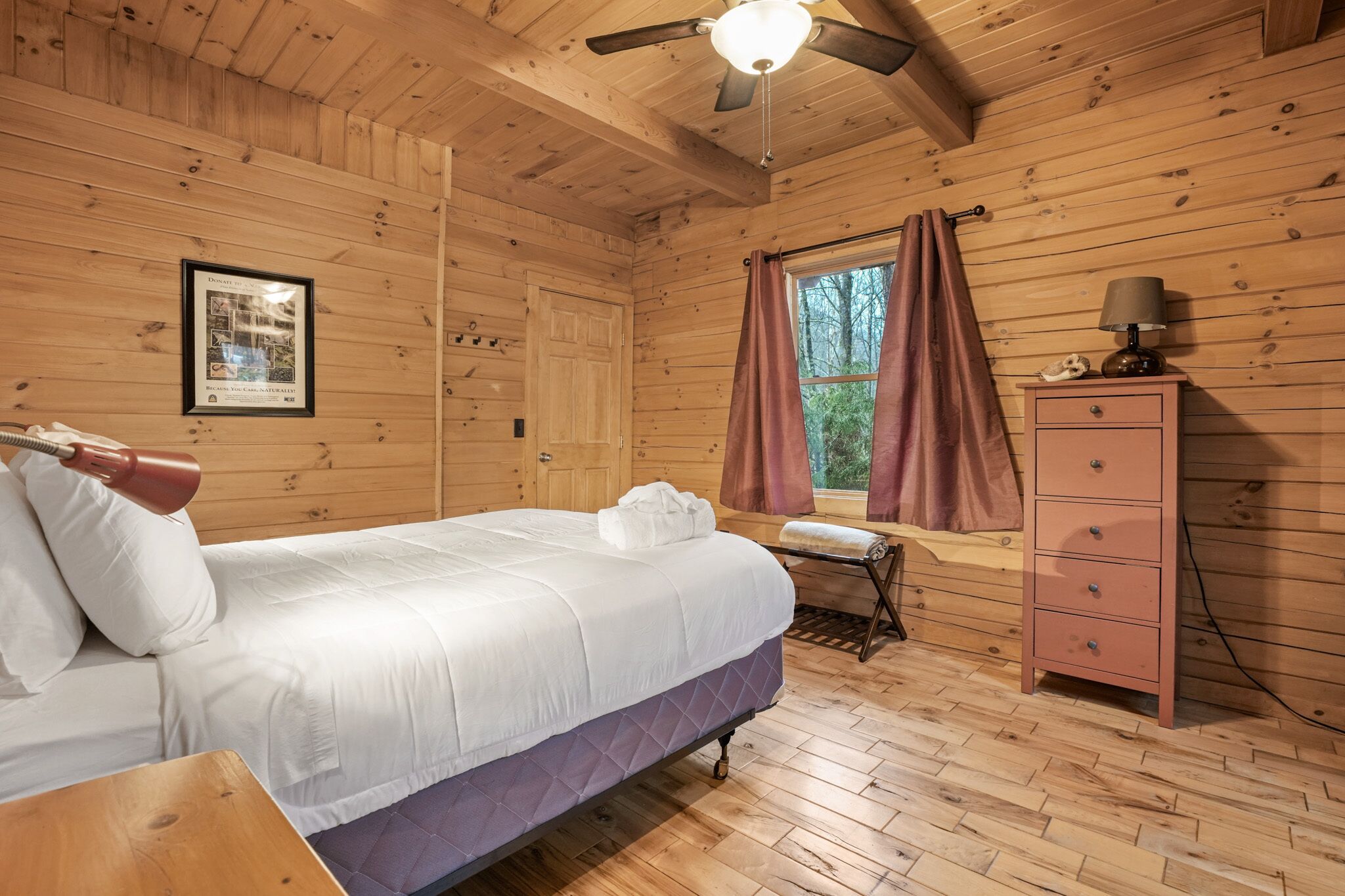 Ein Schlafzimmer einer Holzhütte mit einem Bett, einer Kommode, einem Fenster mit Vorhängen und einem Deckenventilator.