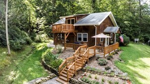 Eine zweistöckige Blockhütte mit einer Terrasse und Sitzbereich im Freien, umgeben von einem gepflegten Garten und Waldgebiet.