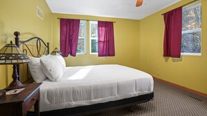 Une chambre à coucher comprenant un lit, une table de chevet, une lampe, un ventilateur de plafond et deux fenêtres avec des rideaux.