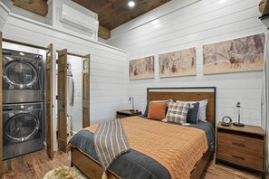 A bedroom with a bed, wooden headboard, and a nightstand with a lamp. There is a washing machine in the adjacent room.