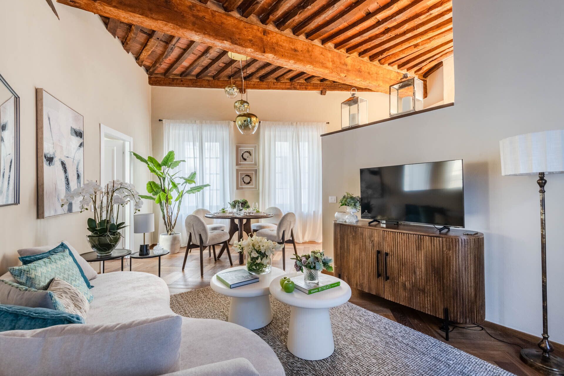 Una sala de estar moderna con techo de madera, televisor de pantalla plana, comedor con mesa y sillas, y sofá con cojines decorativos.