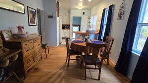 A dining area with a wooden table and chairs, a large teddy bear on a chair, and a kitchen in the background. - Grandma's Cozy 2-bedroom retreat (Muskegon)