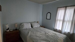 A bedroom with a bed, a nightstand, a lamp, and a window with curtains. - Grandma's Cozy 2-bedroom retreat (Muskegon)