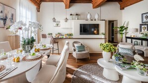 A spacious living room with a wooden ceiling, a dining area with a table set for four, and a kitchen area with a TV and decorative items. - The Mosaic House in Lucca (Lucca)