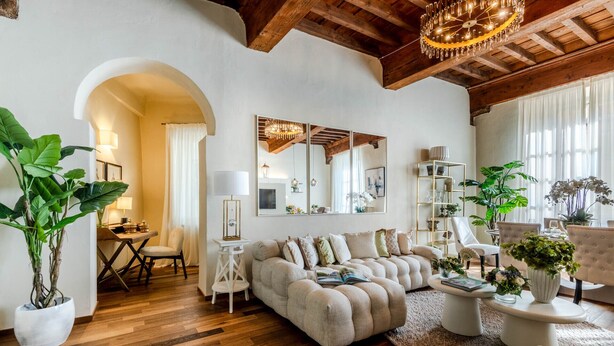 A spacious living room with a large sectional sofa, a wooden floor, and a high ceiling with exposed beams. There is a dining area with a table and chairs, and a large mirror on the wall. - The Mosaic House in Lucca (Lucca)