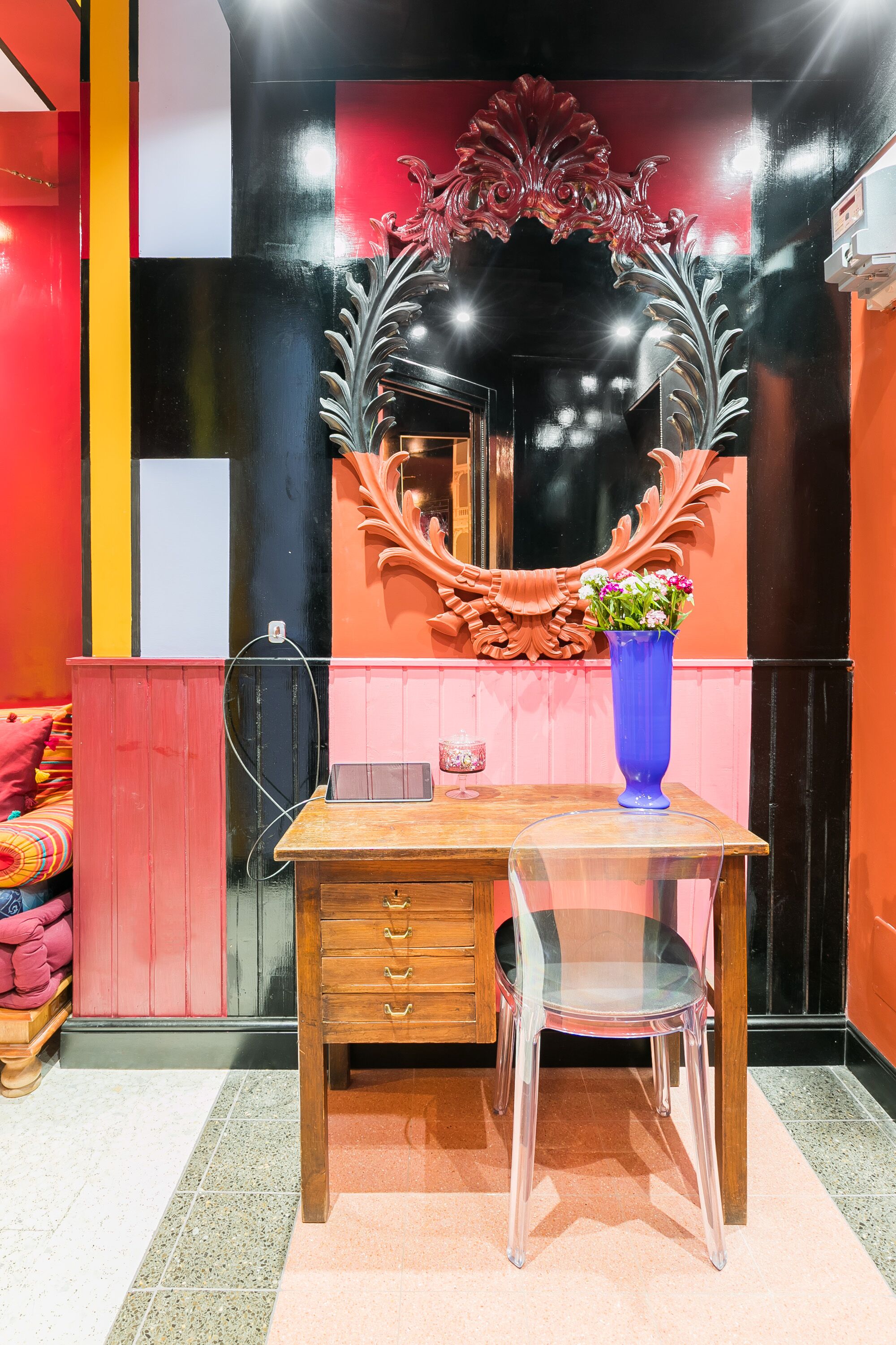A room with a wooden desk, a clear glass chair, a blue vase with flowers, and a decorative mirror with a red and black frame.