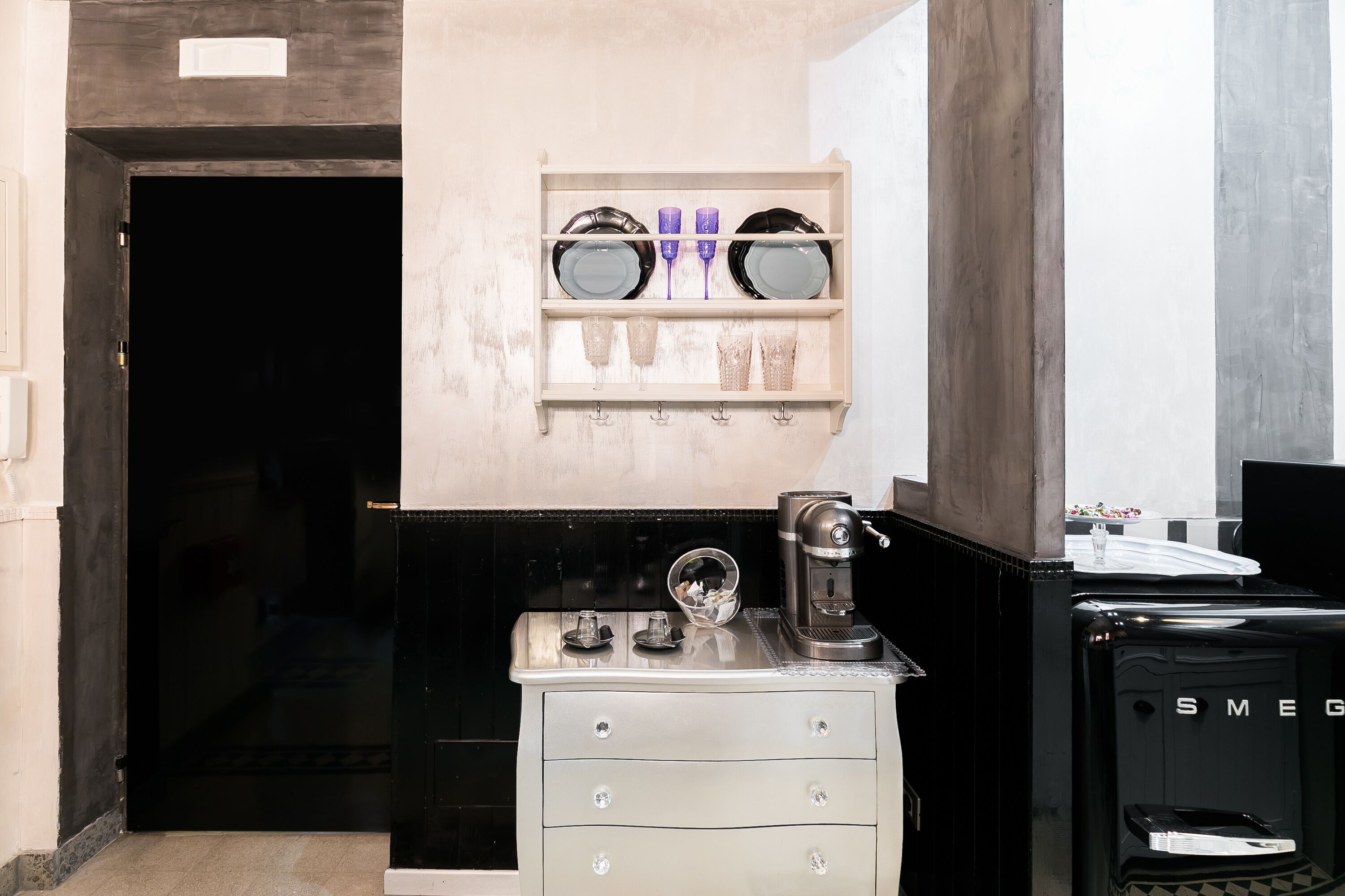 A kitchen with a coffee machine, a black refrigerator, and a shelf with dishes.