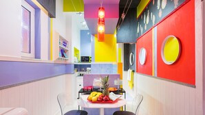Une cuisine aux couleurs vives, avec un coin repas comprenant une table ornée de fruits et d’un ananas, deux chaises et un mur rouge décoré de motifs circulaires.