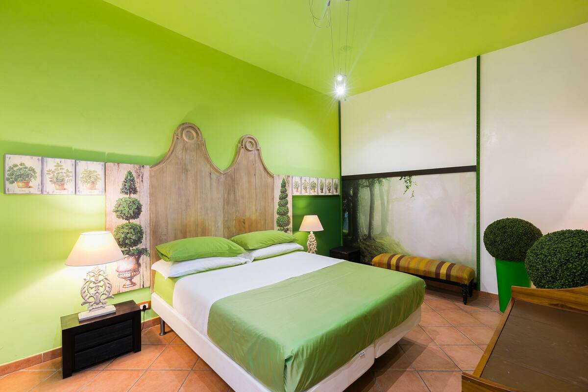 Una habitación con cama, paredes verdes, cabecera de madera, mesita de noche con lámpara y un banco.
