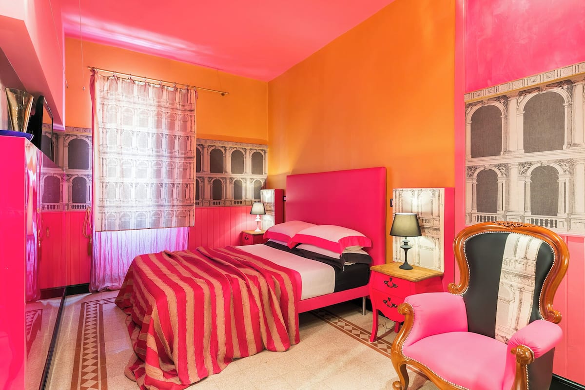 Una habitación con cama, una silla, una mesita de noche y un ventanal con cortinas.