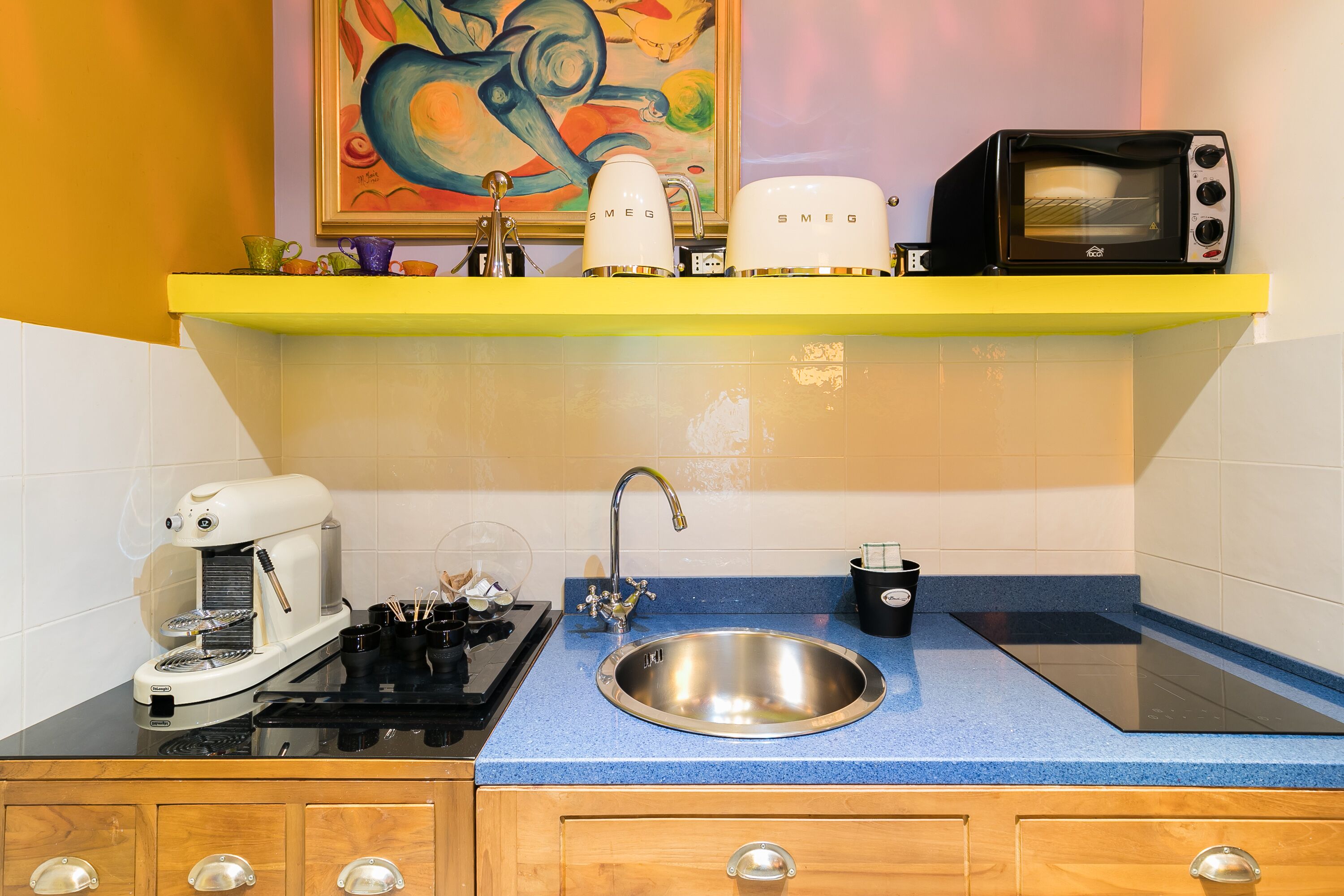 Una cocina con encimera azul, pileta de acero inoxidable, cafetera, tostadora y un cuadro en la pared.
