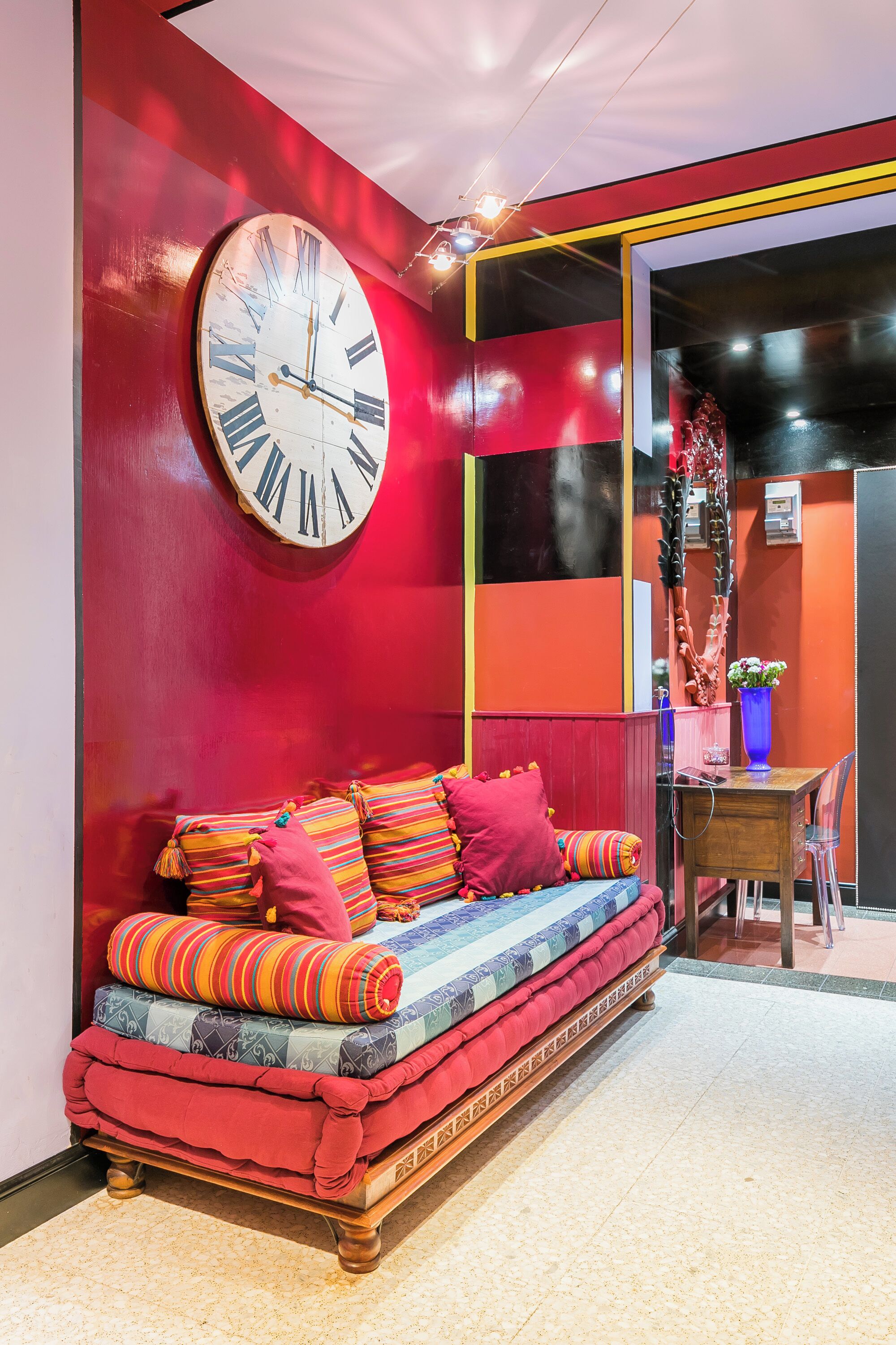 A vibrant room with a large wall clock, a sofa with colorful cushions, and a desk with a vase of flowers.