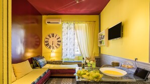 Une pièce avec un mur jaune, un lit avec une tête de lit rouge, une grande horloge murale, une télévision et une table sur laquelle se trouvent des fruits et un saladier.