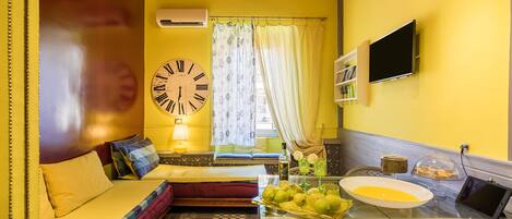Habitación con una pared amarilla, una cama con cabecera roja, un reloj de pared grande, un televisor y una mesa con fruta y un cuenco.