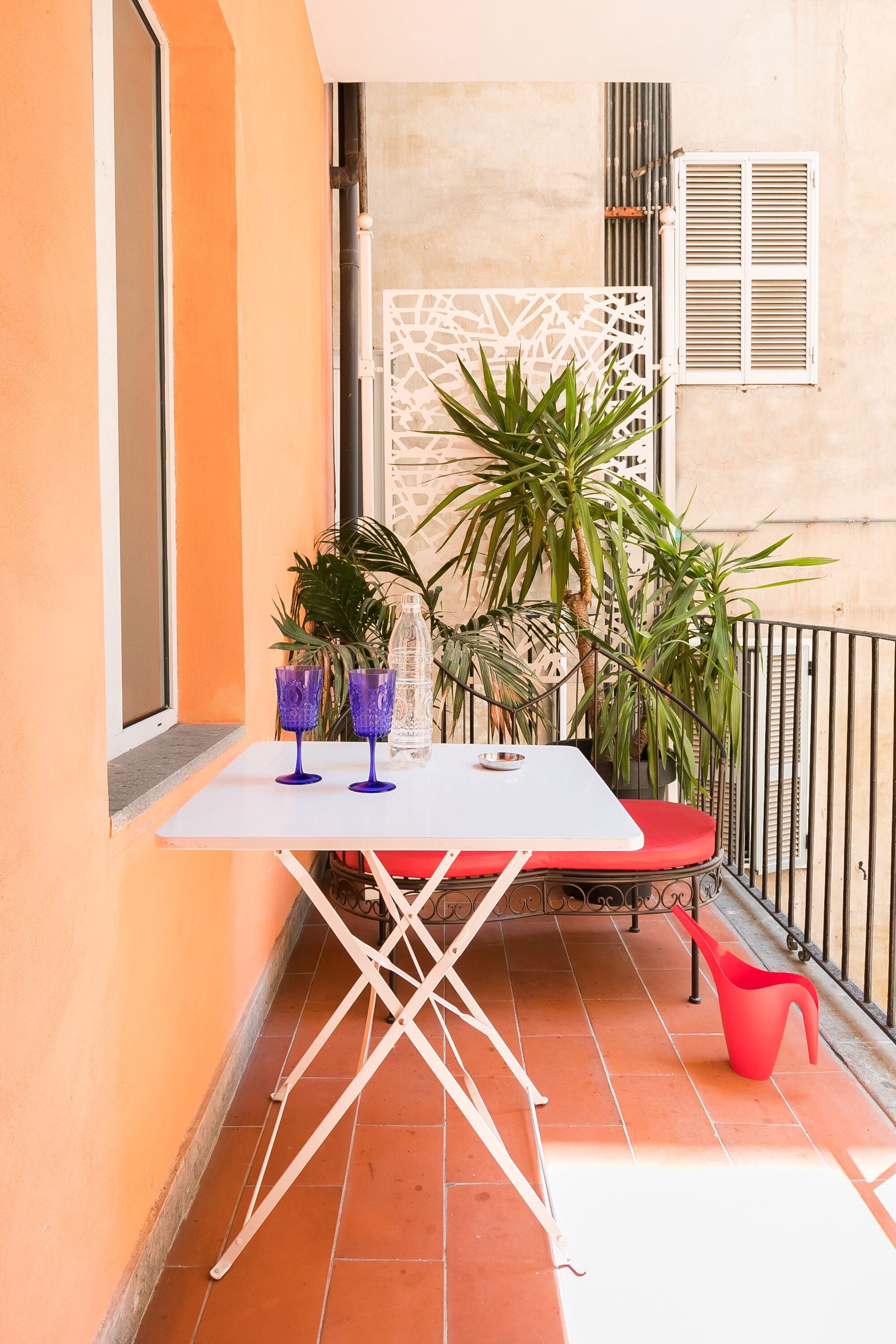 A small outdoor table with two purple glasses and a water bottle, a red chair, and a potted plant on a balcony with orange walls and a metal railing.