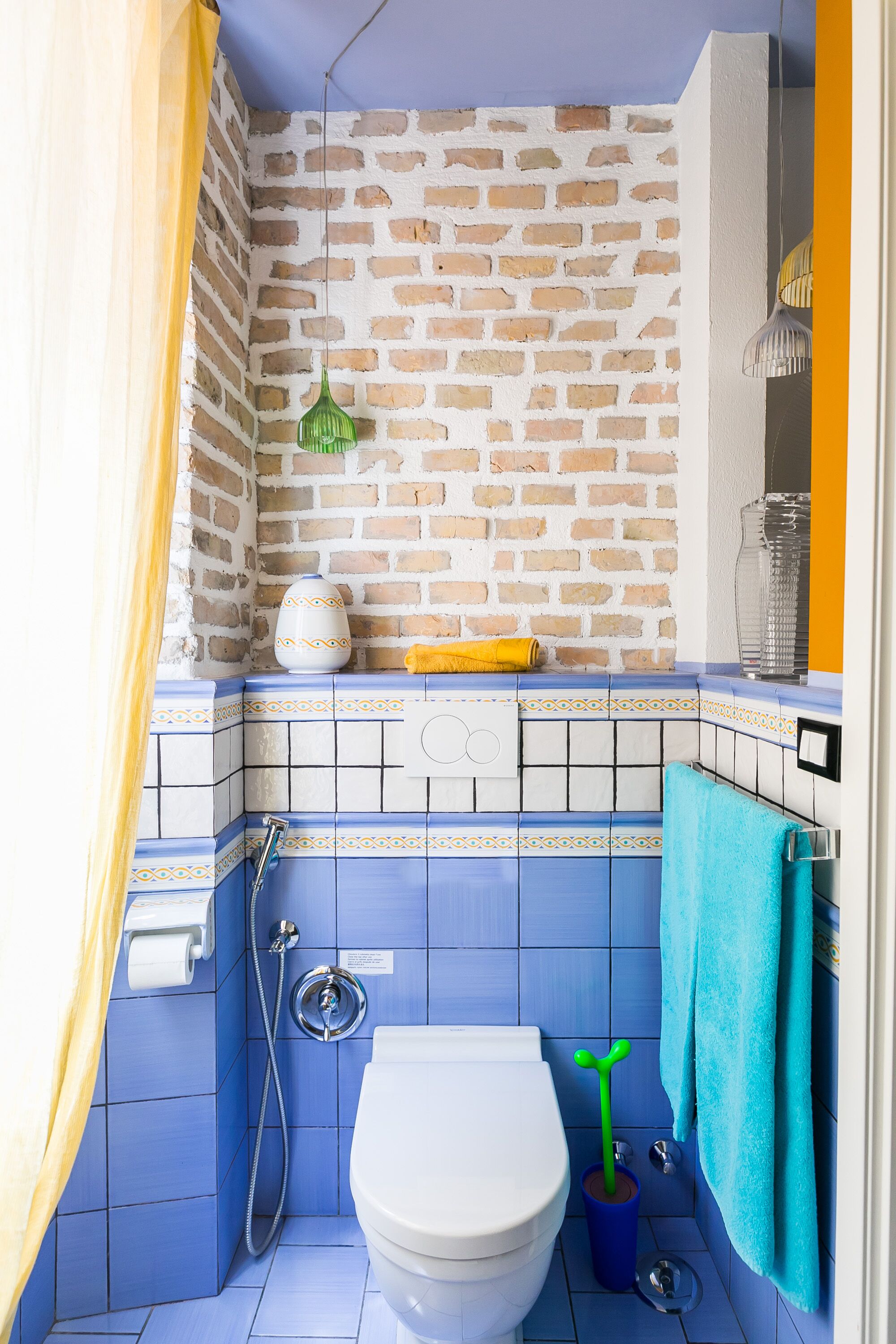 Une salle de bain avec des toilettes blanches, des carreaux bleus, un mur en briques, une serviette jaune et une plante verte.