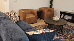 A modern living room with a grey sectional sofa, two brown armchairs, a small round coffee table with a bowl of cookies, and a flat-screen TV mounted on the wall. - Stylish 4BR Home, perfect for families, professionals, business & group stays. (Sherman)