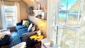 A cozy room with a wooden ceiling, a large window with a view of a lake, a sofa, a small table with a lamp, and a television. - Cozy CABIN on a PRIVATE LAKE in Cabot, AR (Cabot)