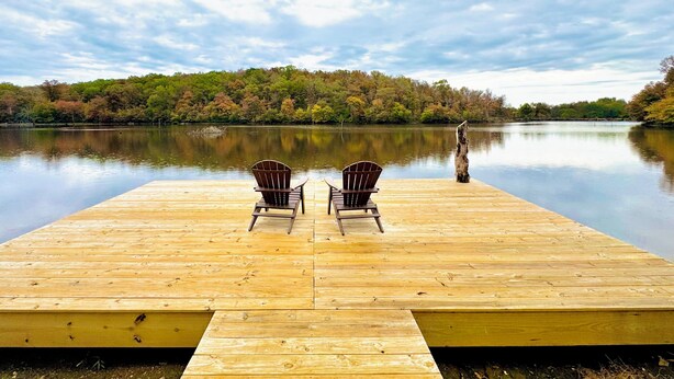 A wooden dock with two Adirondack chairs facing a calm lake, surrounded by trees with autumn foliage. - Cozy CABIN on a PRIVATE LAKE in Cabot, AR (Cabot)