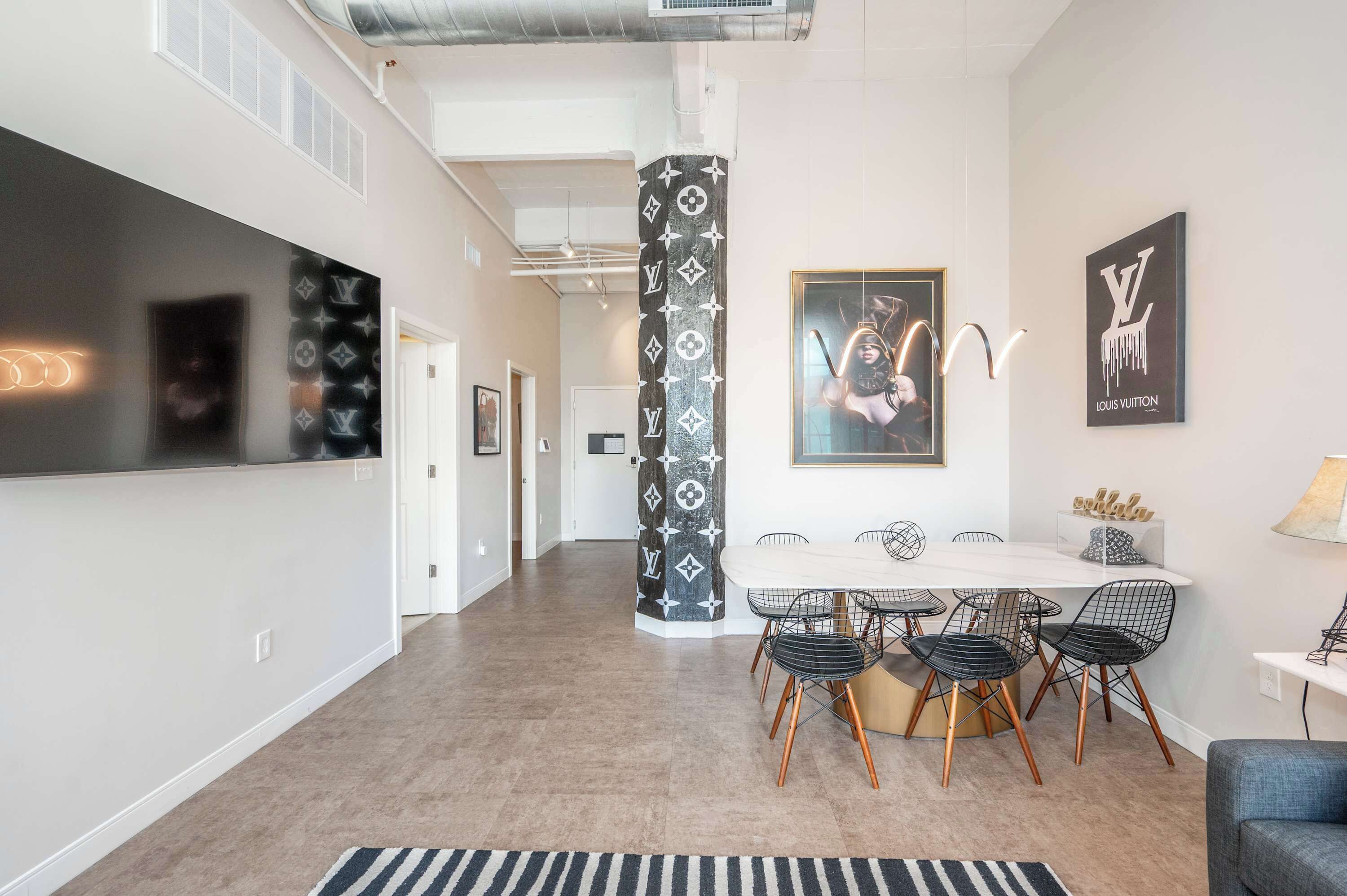Un intérieur moderne avec un coin repas comprenant une table ronde et des chaises en fil de fer, une colonne à motifs noir et blanc, et le logo Louis Vuitton sur le mur.