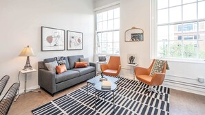 A modern living room with a grey sofa, orange armchairs, a coffee table, and a black and white patterned rug. - The 333 Suites 301 - Suite for 6 (Cincinnati)