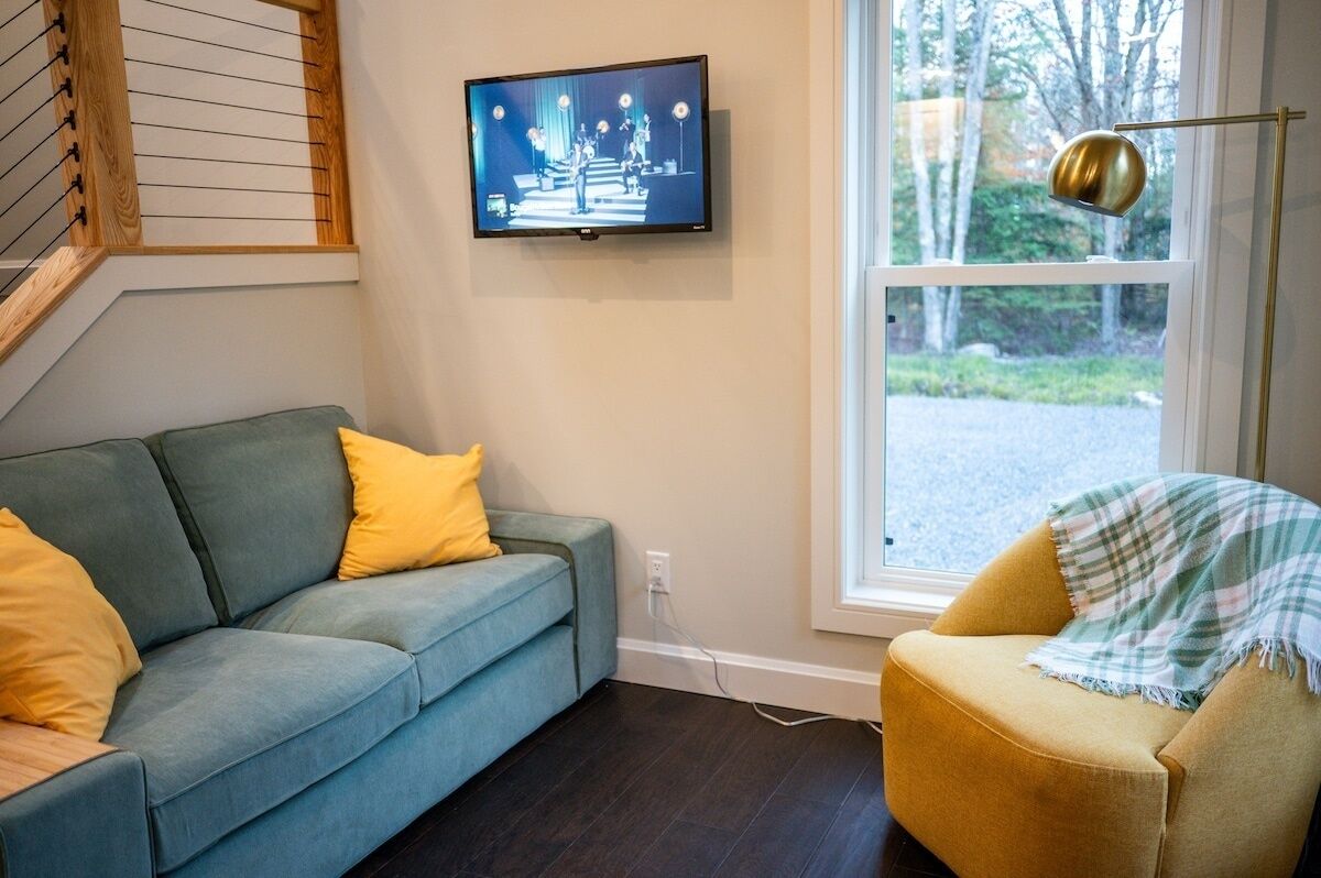 A living room with a blue sofa, a yellow armchair, a television mounted on the wall, and a window with a view of trees.