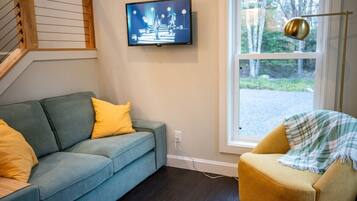 A living room with a blue sofa, a yellow armchair, a television mounted on the wall, and a window with a view of trees.