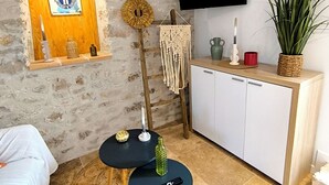 A compact living space with a stone wall, a wooden shelf with a TV, a macrame wall hanging, a small table with a remote, and a cabinet with a plant. - Casa Liã Guest House (Septfonds)