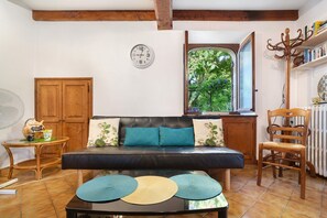 Ein Zimmer mit einer Couch, einem Tisch mit Platzsets, einem Holzstuhl, einem Kleiderständer und einem Fenster mit Blick ins Grüne.