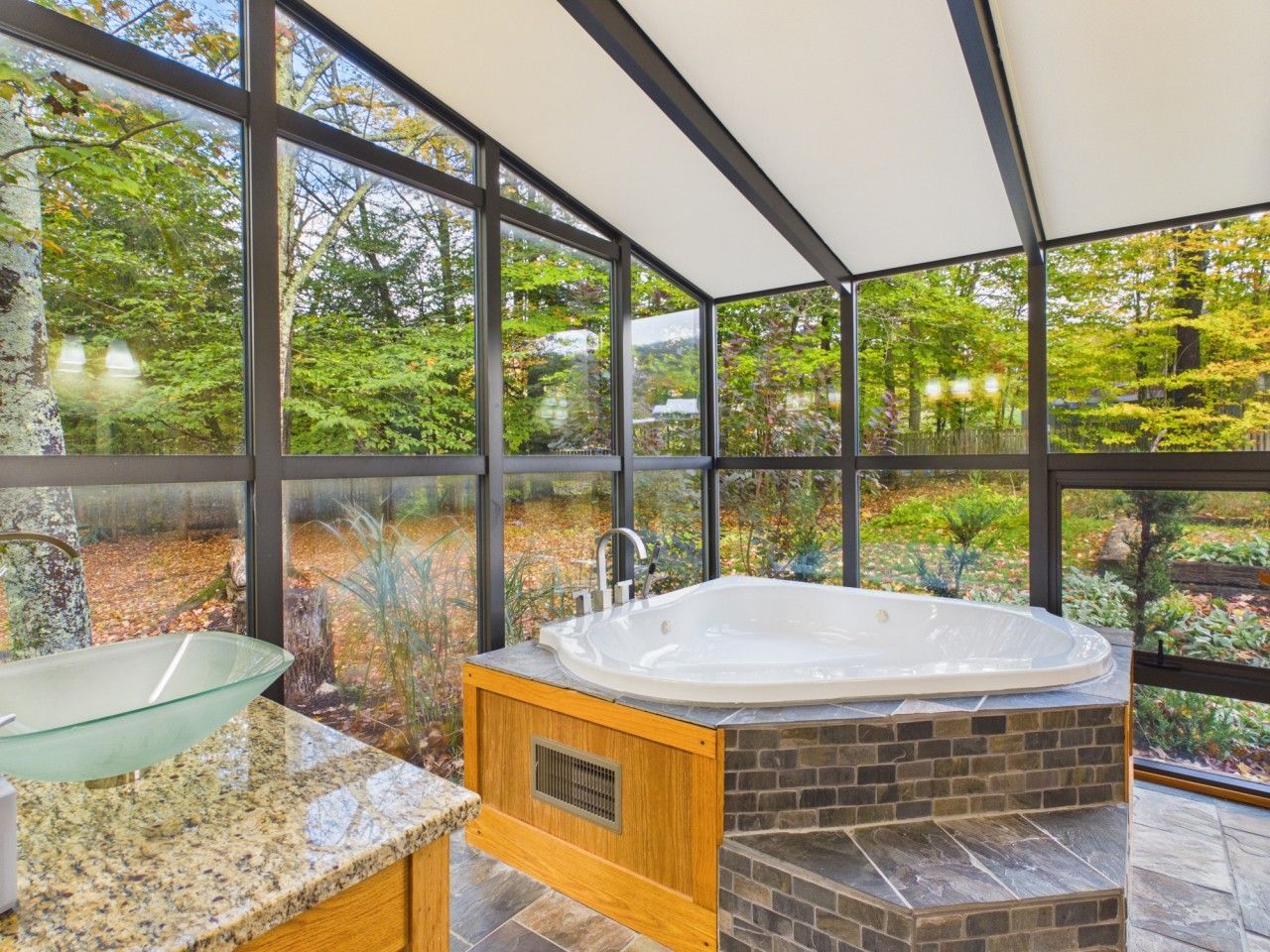 A modern bathroom with a glass sink, a freestanding bathtub, and a view of a lush garden through large windows.