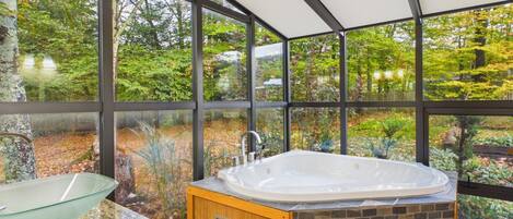 A modern bathroom with a glass sink, a freestanding bathtub, and a view of a lush garden through large windows.