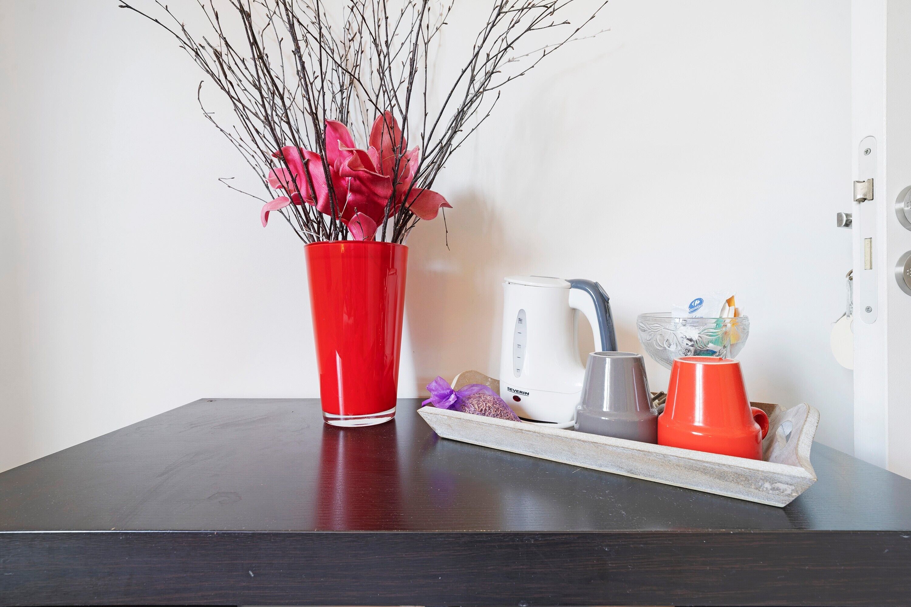 Une table en bois foncé sur laquelle se trouve un vase rouge contenant des branches et des fleurs roses, une bouilloire blanche, une tasse grise et un récipient rouge avec une cuillère.