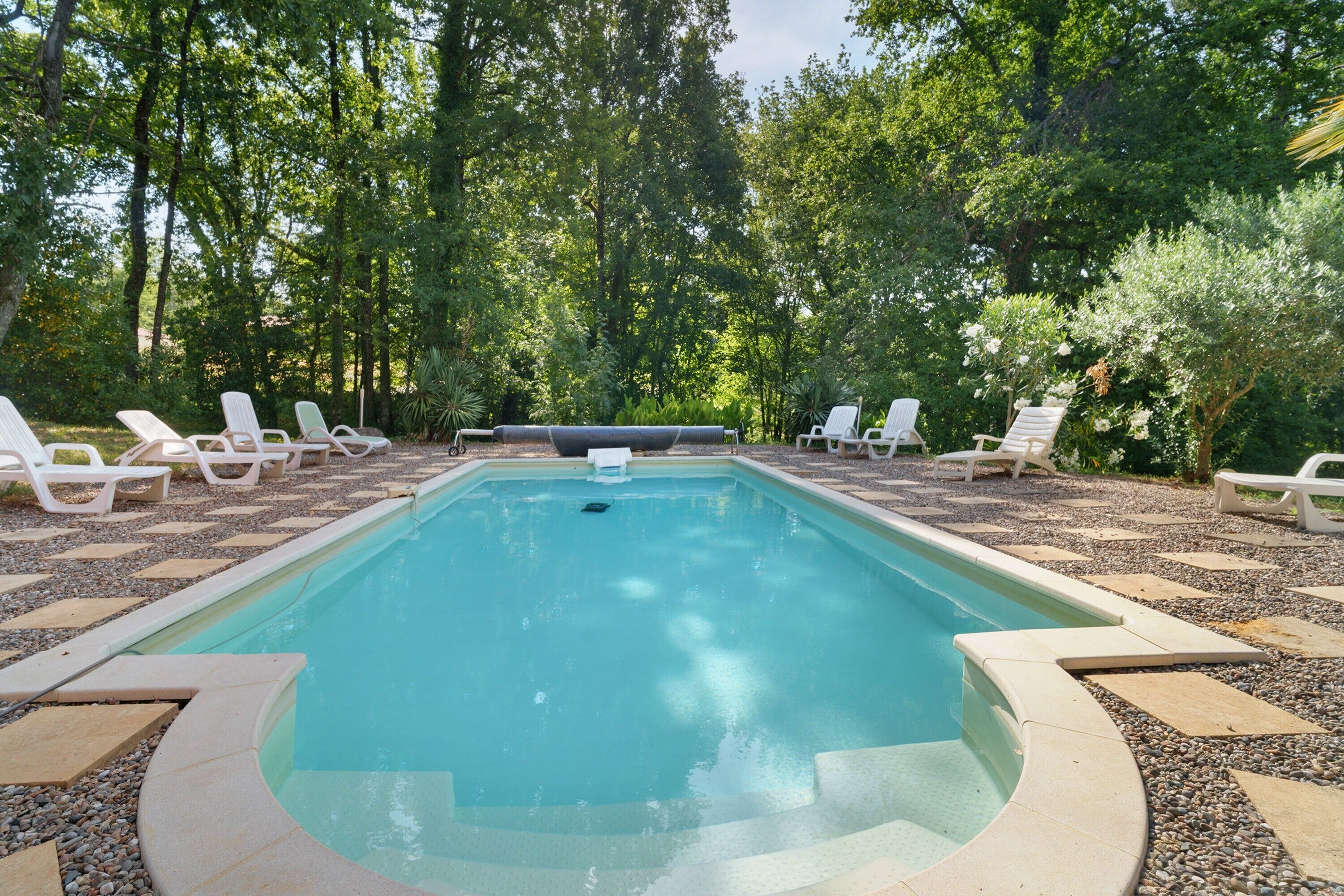 Una piscina rectangular con un jacuzzi en el centro, rodeada de sillones de descanso blancos y un deck con diseño de guijarros.